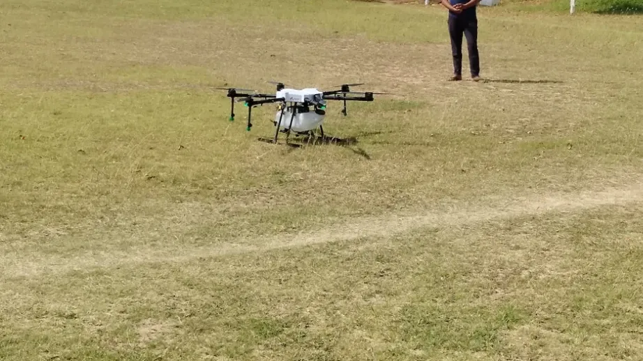 A man wearing a traditional headscarf operates an agricultural drone in an open field at IIVR, Varanasi. The drone is positioned on the ground, ready for use, with trees and buildings in the background and help in innovations in agriculture.